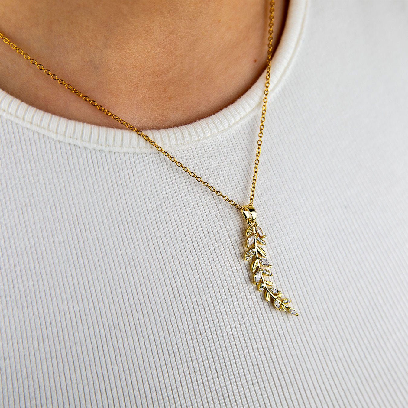 A close-up of a person wearing an 18k gold plated Dahlia Bloom Necklace, featuring a delicate leaf-like pendant adorned with sparkling clear crystals, resting gracefully on a white ribbed shirt.
