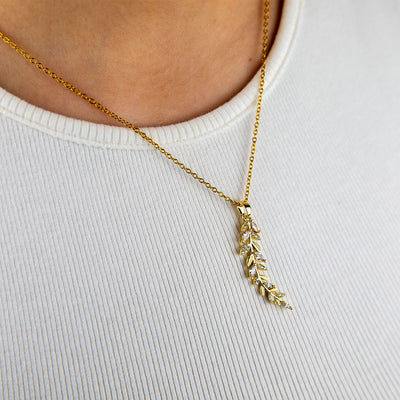 A close-up of a person wearing an 18k gold plated Dahlia Bloom Necklace, featuring a delicate leaf-like pendant adorned with sparkling clear crystals, resting gracefully on a white ribbed shirt.
