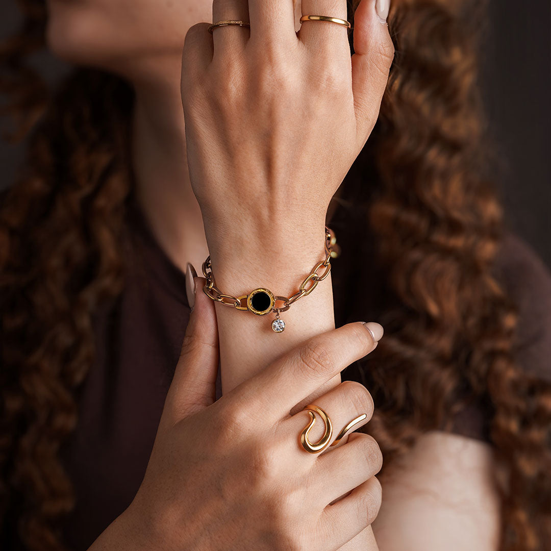A close-up shot of a person's hands and arms, showcasing a variety of gold-toned jewelry. On the wrist is a chunky chain bracelet with a black and gold circular charm from which a small gemstone hangs. The fingers are adorned with several rings: a simple thin band, a thicker polished band, and a bold, wave-like ring.