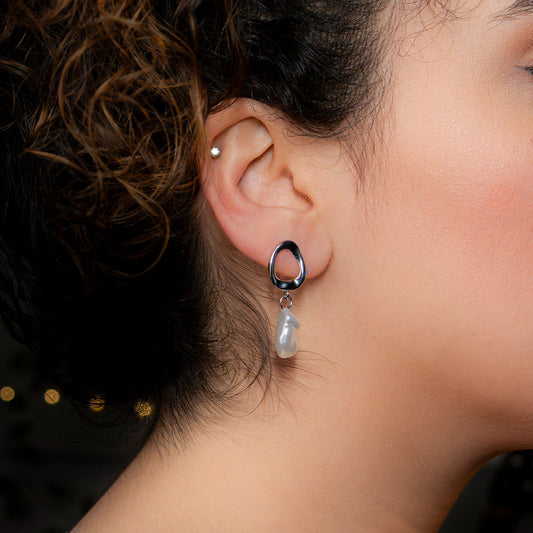 Close-up of a person's ear wearing a dangle earring. The earring features a polished, tear-drop shaped gold loop from which a single, irregular-shaped white pearl hangs. The earring is called Alba Teardrop Pearl Drops.