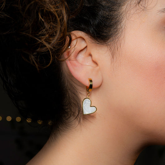 A close-up side profile shows a person's ear adorned with a gold hoop earring. Dangling from the hoop is a white, iridescent heart-shaped charm with a gold border. This lovely piece is known as the Venus Heart Drops.