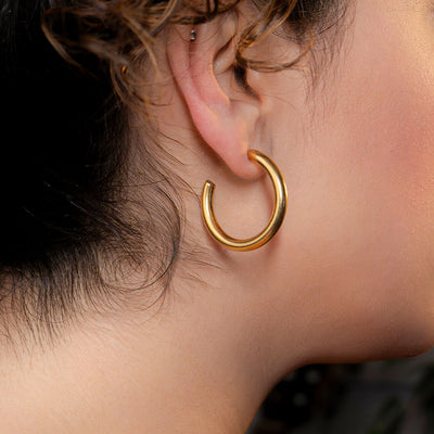 A close-up side profile shot of a person's ear, showcasing a large, thick, and perfectly circular gold hoop earring. The earring has a very shiny, polished surface. This impactful piece is named the Marina Bold Hoops.
