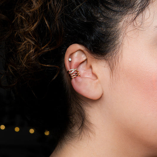 A close-up side profile of a person's ear, showcasing a gold ear cuff that wraps around the conch. The ear cuff consists of multiple thin bands; some are plain gold, while others are encrusted with small, clear gemstones. This radiant accessory is called the Nova Sparkle Ear Cuff.