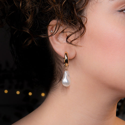 A close-up side profile of a person's ear, featuring a polished gold hoop earring from which a large, irregularly shaped white baroque pearl hangs. This elegant piece is named the Aurelia Pearl Drop Hoops.