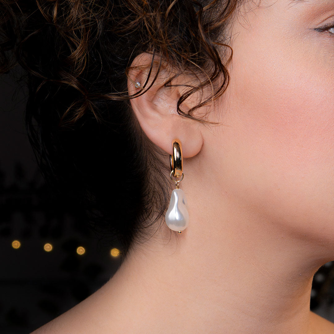 A close-up side profile of a person's ear, featuring a polished gold hoop earring from which a large, irregularly shaped white baroque pearl hangs. This elegant piece is named the Aurelia Pearl Drop Hoops.