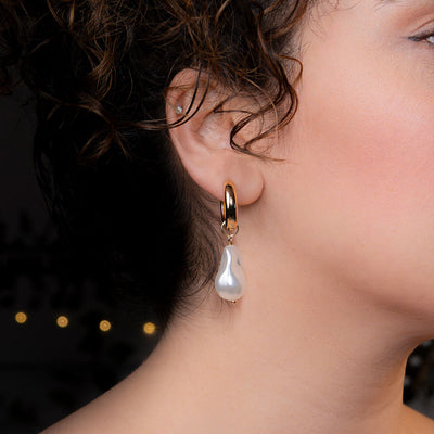 A close-up side profile of a person's ear, featuring a polished gold hoop earring from which a large, irregularly shaped white baroque pearl hangs. This elegant piece is named the Aurelia Pearl Drop Hoops.