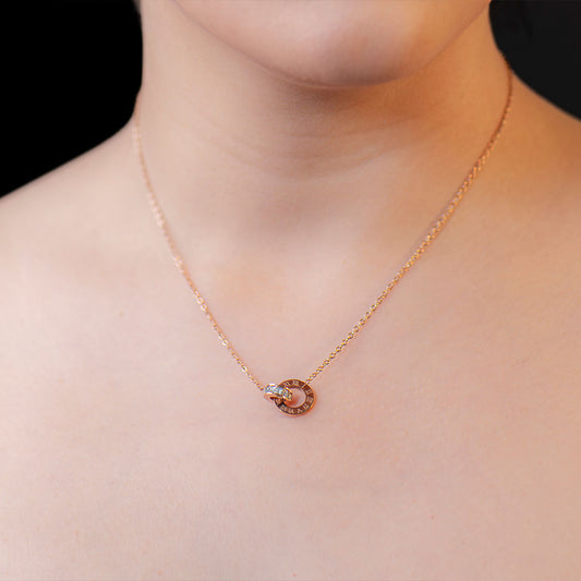A close-up shot of a person's bare chest and neck, showcasing a delicate gold chain necklace with a circular pendant. The pendant consists of two interlocking rings; one ring has Roman numerals engraved on it, and the other is adorned with a single small, clear gemstone. This elegant piece is known as the Olympia Roman Pendant.