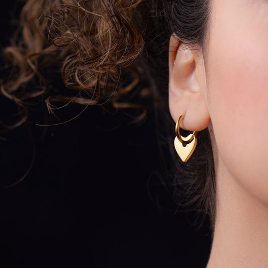 A close-up side profile of a person's ear, showcasing a small, shiny gold hoop earring with a solid, polished gold heart-shaped lock charm dangling from it. This endearing piece is called the Cora Love Hoops.
