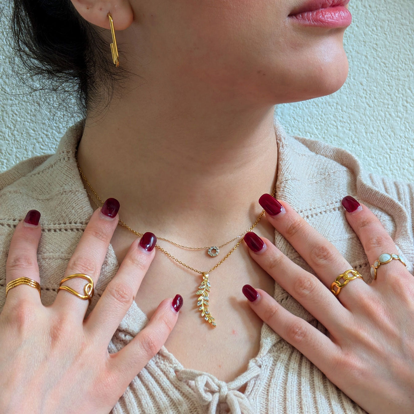 A close-up of a person adorned with a luxurious collection of 18k gold plated jewelry. This includes a Dahlia Bloom Necklace with a delicate leaf-shaped pendant, complemented by a layered delicate chain. On her fingers, a Hazel Spark Ring, Isolde Luxe Ring, Seraphine Charm Ring, and Delphine Shine Ring are elegantly displayed, all against a neutral-toned top.