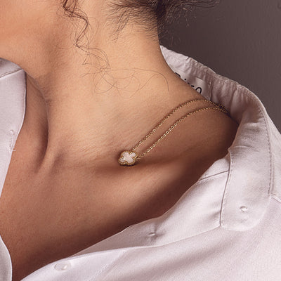 A close-up of a person's neck and shoulder, showcasing an 18k gold plated Sophia Timeless Clover Necklace with a delicate white clover pendant, resting elegantly on a white collared shirt.