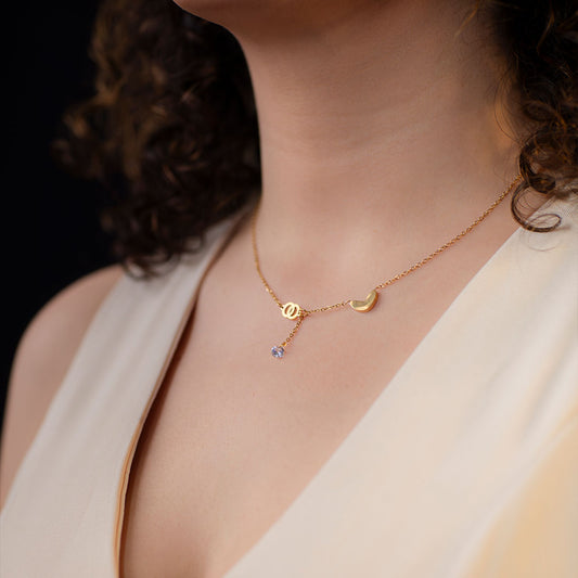 A close-up shot of a person's upper chest and neck, wearing a delicate gold chain necklace. The necklace features an asymmetrical design with a small gold crescent moon charm on one side and a circular gold charm with a small, dangling light purple gemstone on the other. This whimsical piece is called the Luna Whisper Necklace.