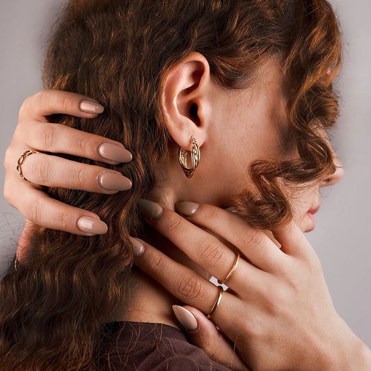 A close-up side profile of a person with curly hair, focusing on their hands and ear. The person is wearing a small, intricately patterned gold hoop earring, a simple gold ring on one finger, and a bolder, textured ring on another.