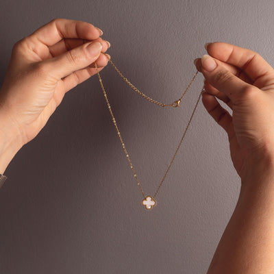 Two hands holding up an 18k gold plated Sophia Timeless Clover Necklace, showcasing its delicate chain and the white clover pendant with a subtle beaded edge against a dark grey background.