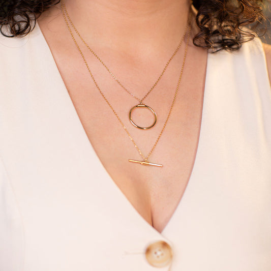 A close-up shot of a person's upper chest and neck, wearing a white v-neck top. They are adorned with a layered gold necklace featuring two distinct pendants: an upper chain with a polished open circle, and a lower chain with a horizontal bar. This elegant set is called the Avani Layered Luxe Necklace.