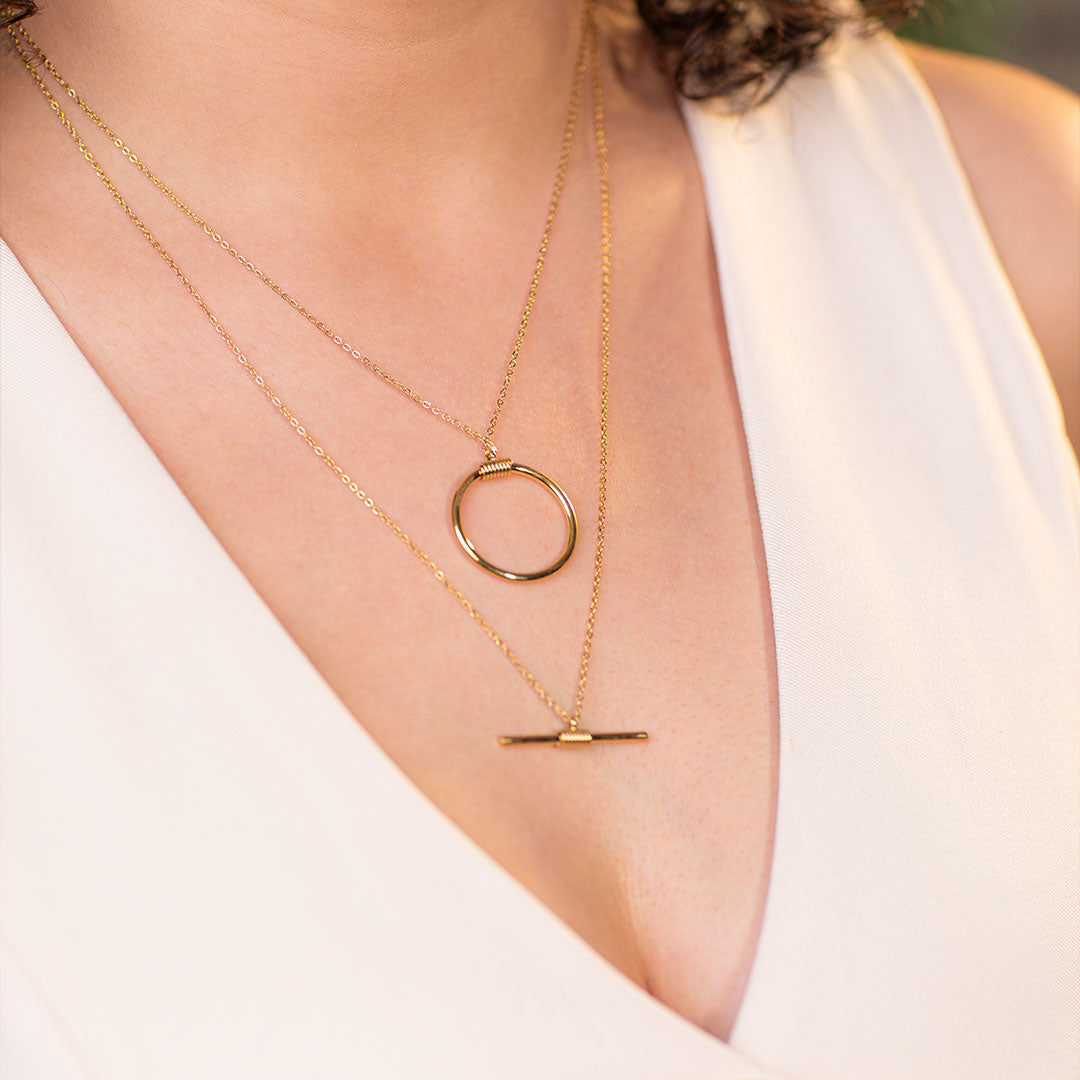 Captured in a close-up, a person wearing a chic white v-neck top proudly displays the Avani Layered Luxe Necklace, a stunning gold piece featuring two layers. The shorter layer boasts a delicate, open circle pendant, while the longer chain carries a sleek, linear bar.