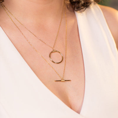 Captured in a close-up, a person wearing a chic white v-neck top proudly displays the Avani Layered Luxe Necklace, a stunning gold piece featuring two layers. The shorter layer boasts a delicate, open circle pendant, while the longer chain carries a sleek, linear bar.