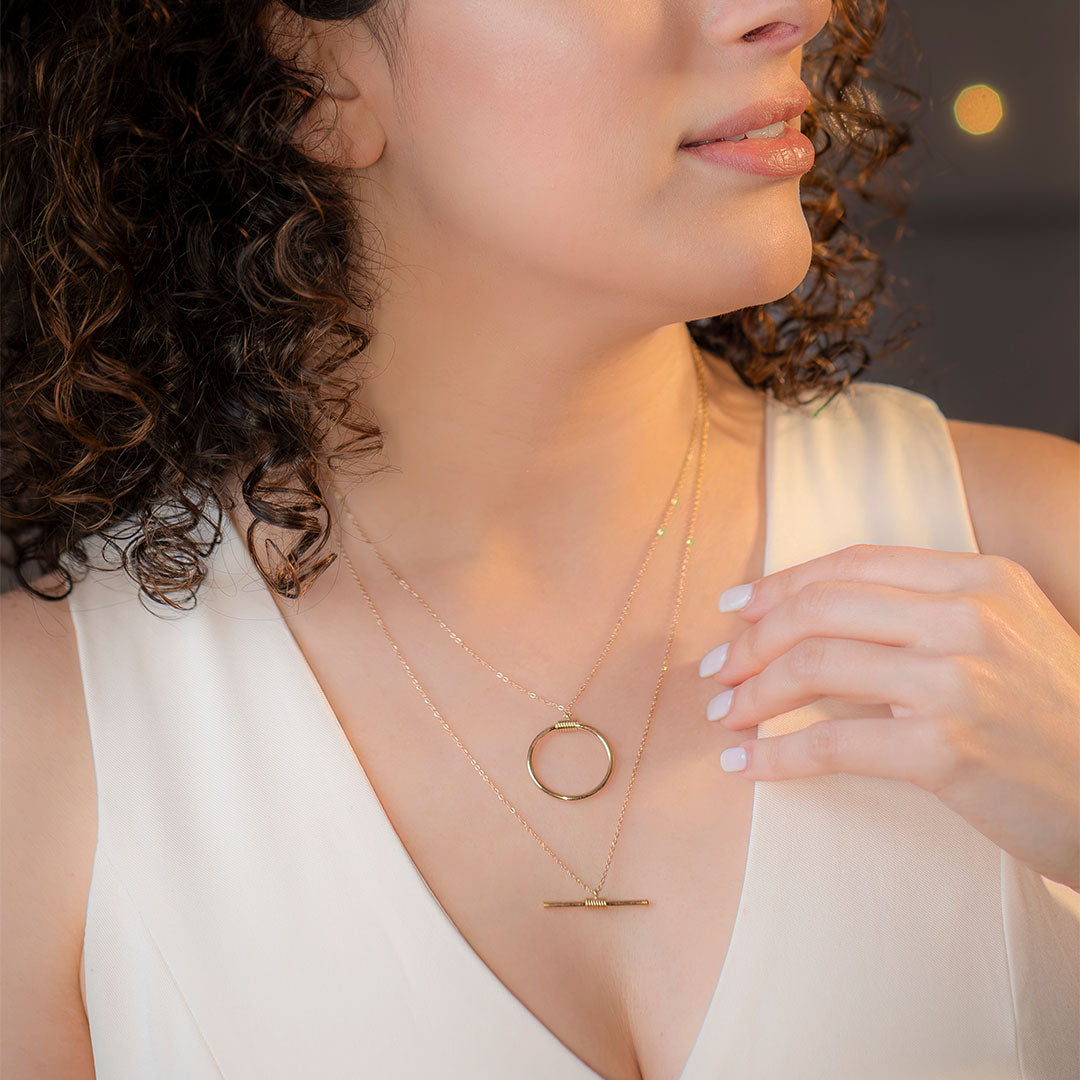 Framing the neckline of a white v-neck top, this close-up image showcases the beautiful Avani Layered Luxe Necklace. This gold-toned layered piece is detailed with an upper chain holding a minimalist circular charm and a lower chain suspending a modern horizontal bar.