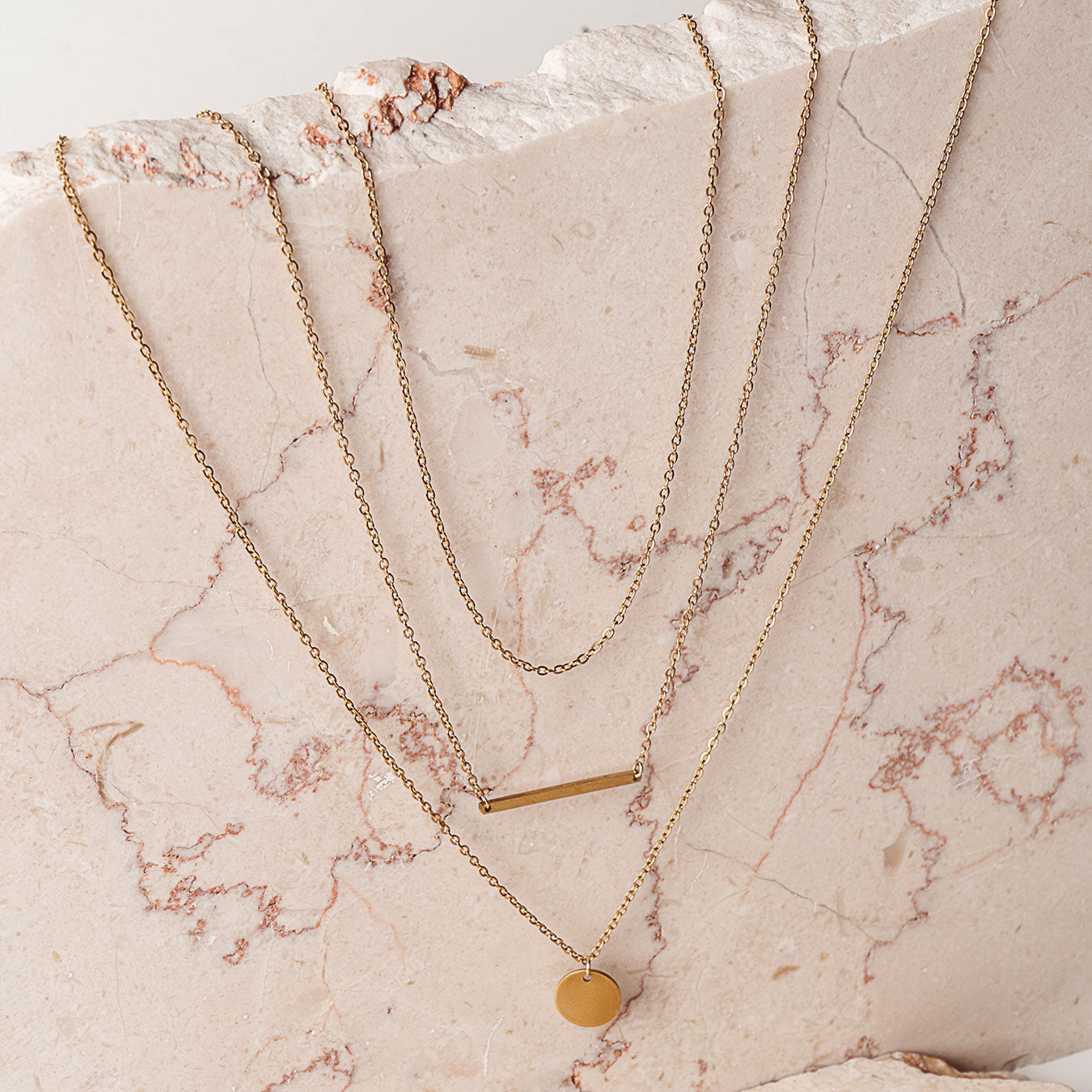 Three elegant 18k gold plated Camellia Classic Chain necklaces are artfully layered on a light-colored, veined marble surface. One chain features a minimalist bar pendant, another a simple circular disc pendant, and the third is a plain chain, demonstrating versatile styling options.