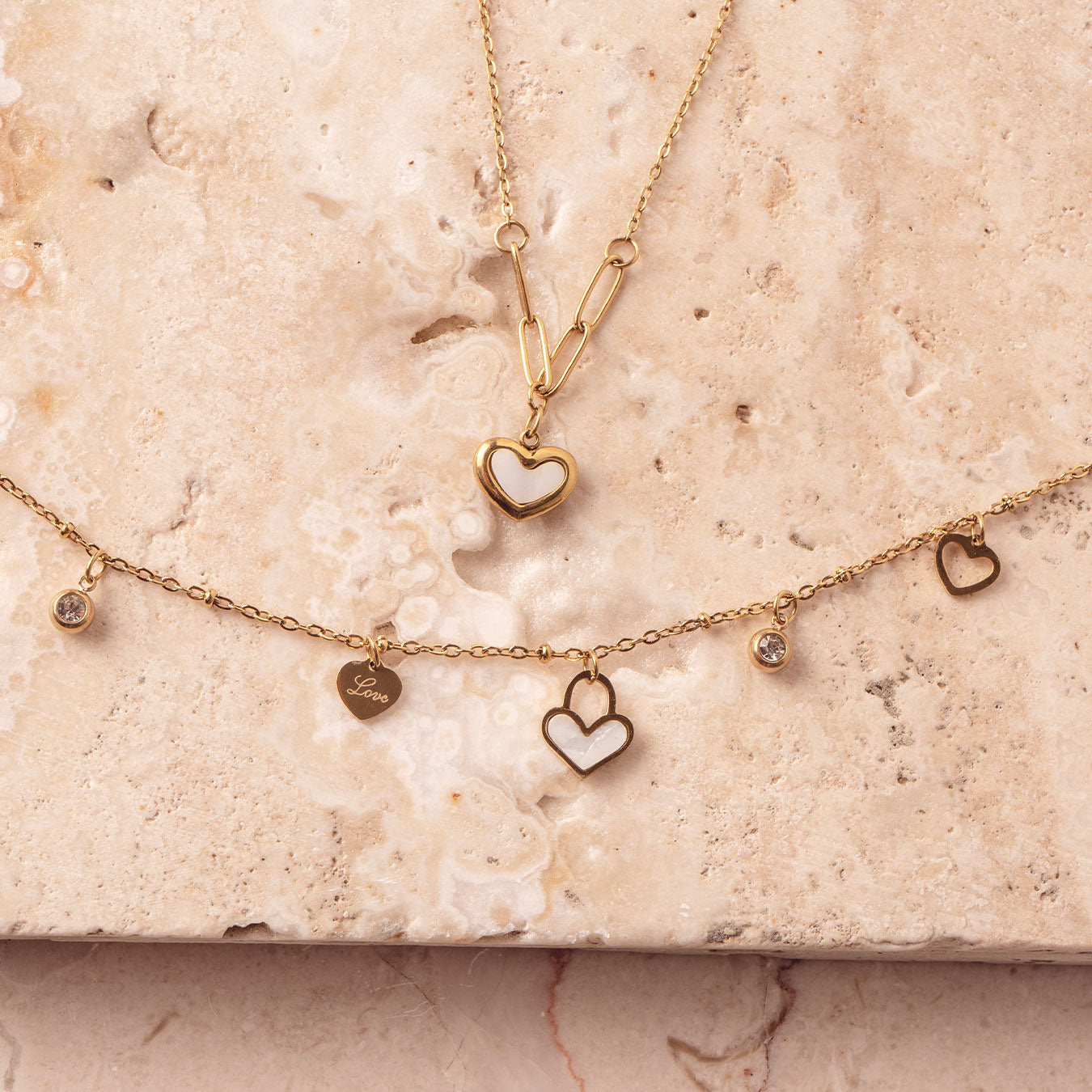 Two delicate 18k gold plated necklaces arranged on a light-colored marble surface. The top necklace features a Violet Petal Pendant, while the bottom necklace showcases multiple Amara Love Charms, including heart-shaped and sparkling designs.