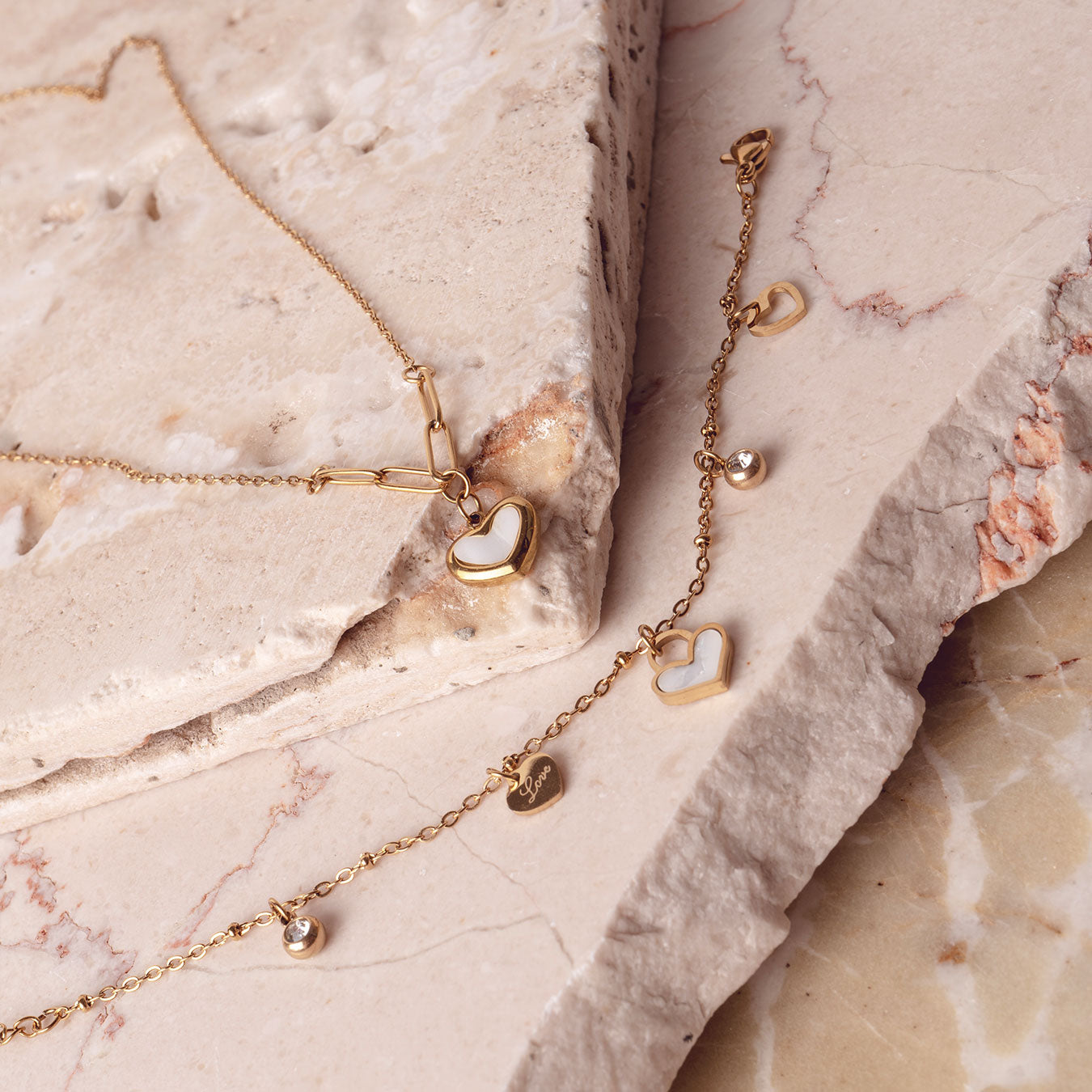 Two elegant 18k gold plated necklaces arranged on a textured stone surface. The top necklace features a Violet Petal Pendant with a white heart charm, while the bottom necklace showcases delicate Amara Love Charms, including small hearts and sparkling details.
