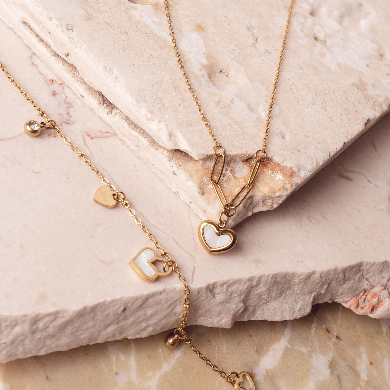 Two stunning 18k gold plated necklaces artfully arranged on a textured light-colored stone surface. The upper necklace features a Violet Petal Pendant with a prominent white heart charm, while the lower necklace showcases multiple delicate Amara Love Charms, including small heart shapes and sparkling accents.