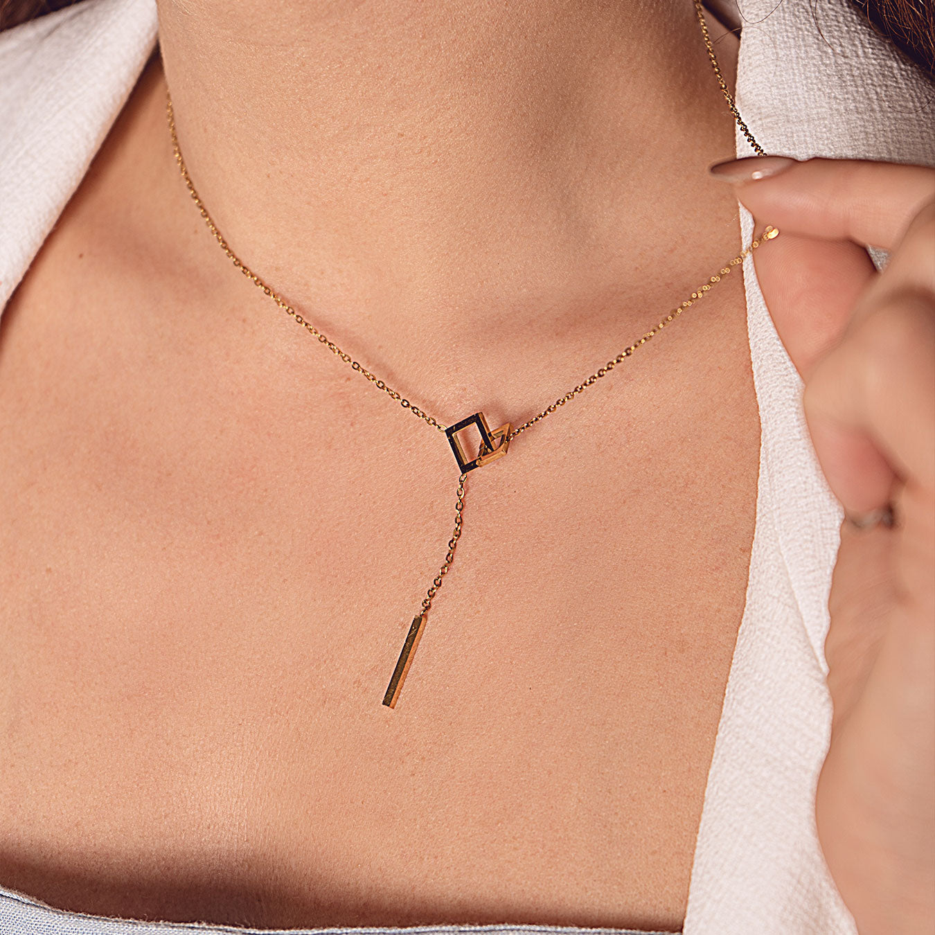 A close-up of a person's neck and upper chest, featuring an elegant 18k gold plated Jasmine Grace Necklace. The necklace showcases a modern lariat design with an open square and a dangling rectangular bar, complementing a light-colored collared shirt.