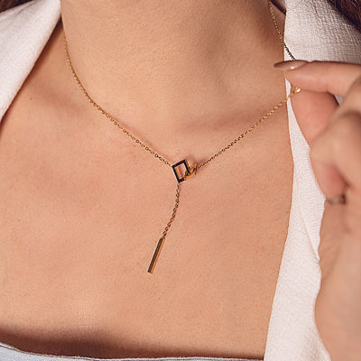 A close-up of a person's neck and upper chest, featuring an elegant 18k gold plated Jasmine Grace Necklace. The necklace showcases a modern lariat design with an open square and a dangling rectangular bar, complementing a light-colored collared shirt.