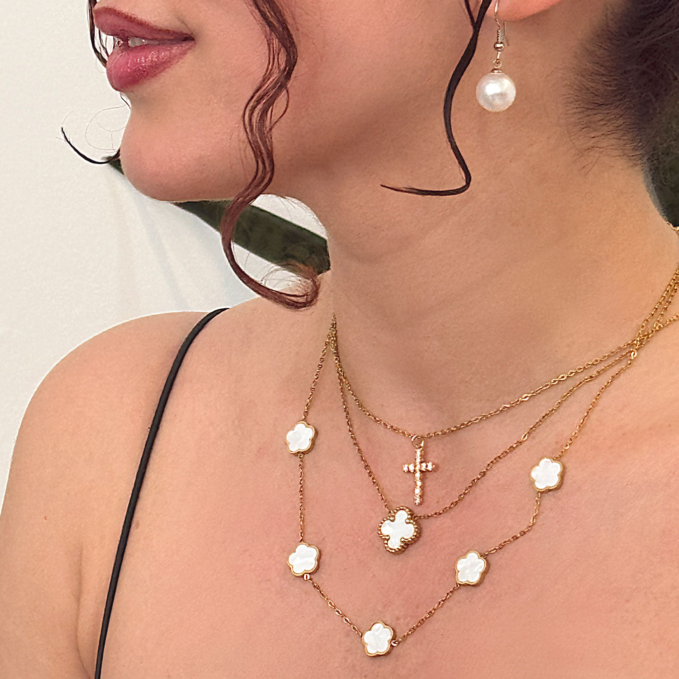 A close-up of a person's neckline adorned with a beautiful layered arrangement of 18k gold plated necklaces. This includes a Luminous Faith Pendant in the shape of a sparkling cross, a Celestia Bloom Necklace with multiple white floral charms, and a Sophia Timeless Clover Necklace, completing a sophisticated look.