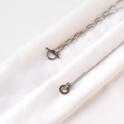 Two silver-toned necklaces gracefully displayed on a soft white fabric. The Lustrous Arc Necklace features a substantial chain with a toggle clasp, while the Aurora Trend Necklace showcases a delicate chain with a sparkling circular pendant adorned with Roman numerals.