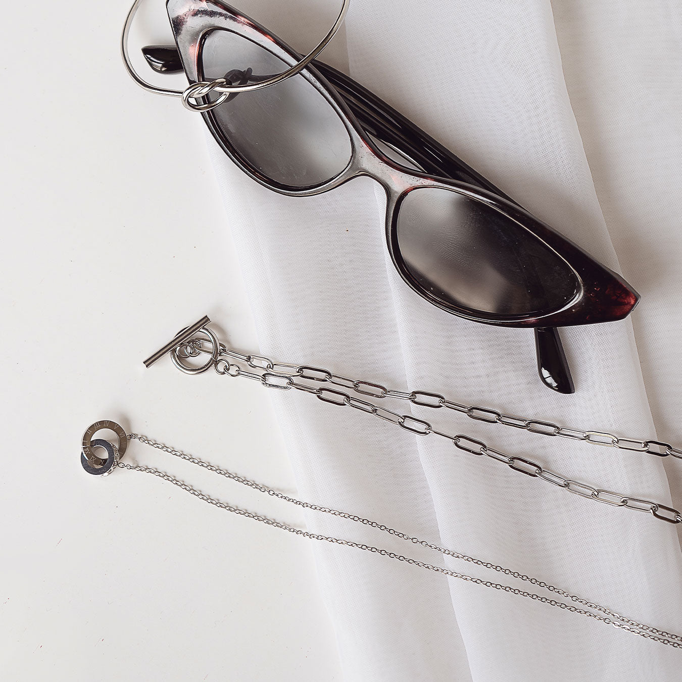 A flat lay arrangement featuring a pair of stylish sunglasses resting on a white fabric. Below them, a silver-toned Lustrous Arc Necklace with a toggle clasp and a delicate Aurora Trend Necklace with a circular Roman numeral charm are elegantly displayed. A Seraphine Gold Bracelet (though appearing silver-toned in this image) is also part of the arrangement, completing a chic accessory collection.