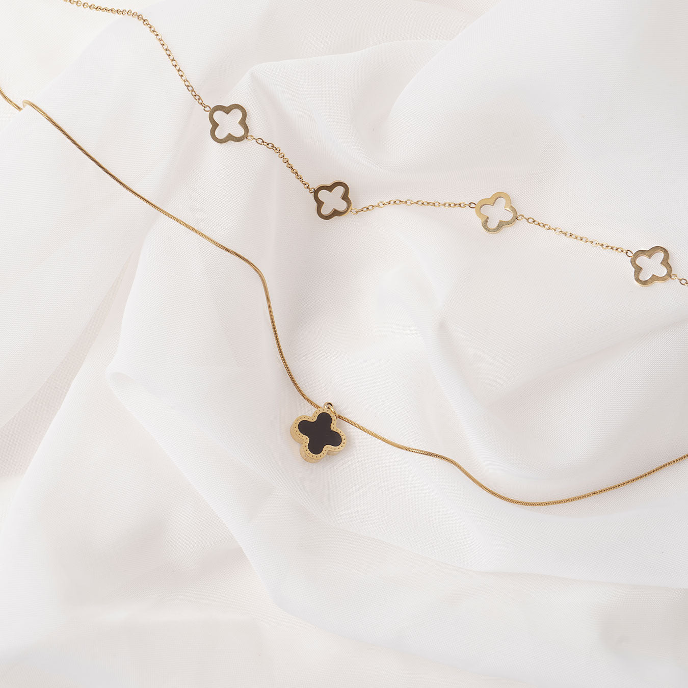 Two elegant 18k gold plated necklaces displayed on a soft, draped white fabric. The lower necklace features a single Serena Clover Pendant with a dark center, while the upper necklace showcases multiple open clover charms.