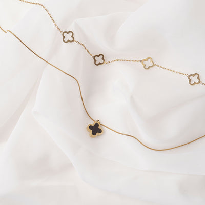 Two elegant 18k gold plated necklaces displayed on a soft, draped white fabric. The lower necklace features a single Serena Clover Pendant with a dark center, while the upper necklace showcases multiple open clover charms.