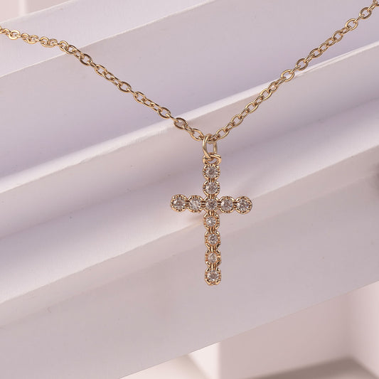 A close-up of an 18k gold plated Luminous Faith Pendant, a sparkling cross necklace adorned with numerous crystals, resting on a clean white ribbed surface.