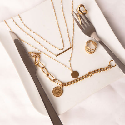 An elegant display of 18k gold plated jewelry presented on a white square plate alongside a knife and fork. The collection includes a Camellia Classic Chain necklace (appearing in a layered fashion with bar and disc pendants), a Lila Eternity Band, a Mila Zircon Ring, and an Isolde Luxe Bangle featuring a coin charm and chain link design.