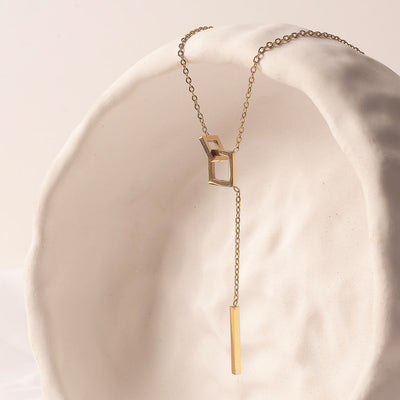An elegant 18k gold plated Jasmine Grace Necklace, featuring a unique lariat design with an open square link and a sleek rectangular bar pendant, gracefully draped over a textured white sculpture.