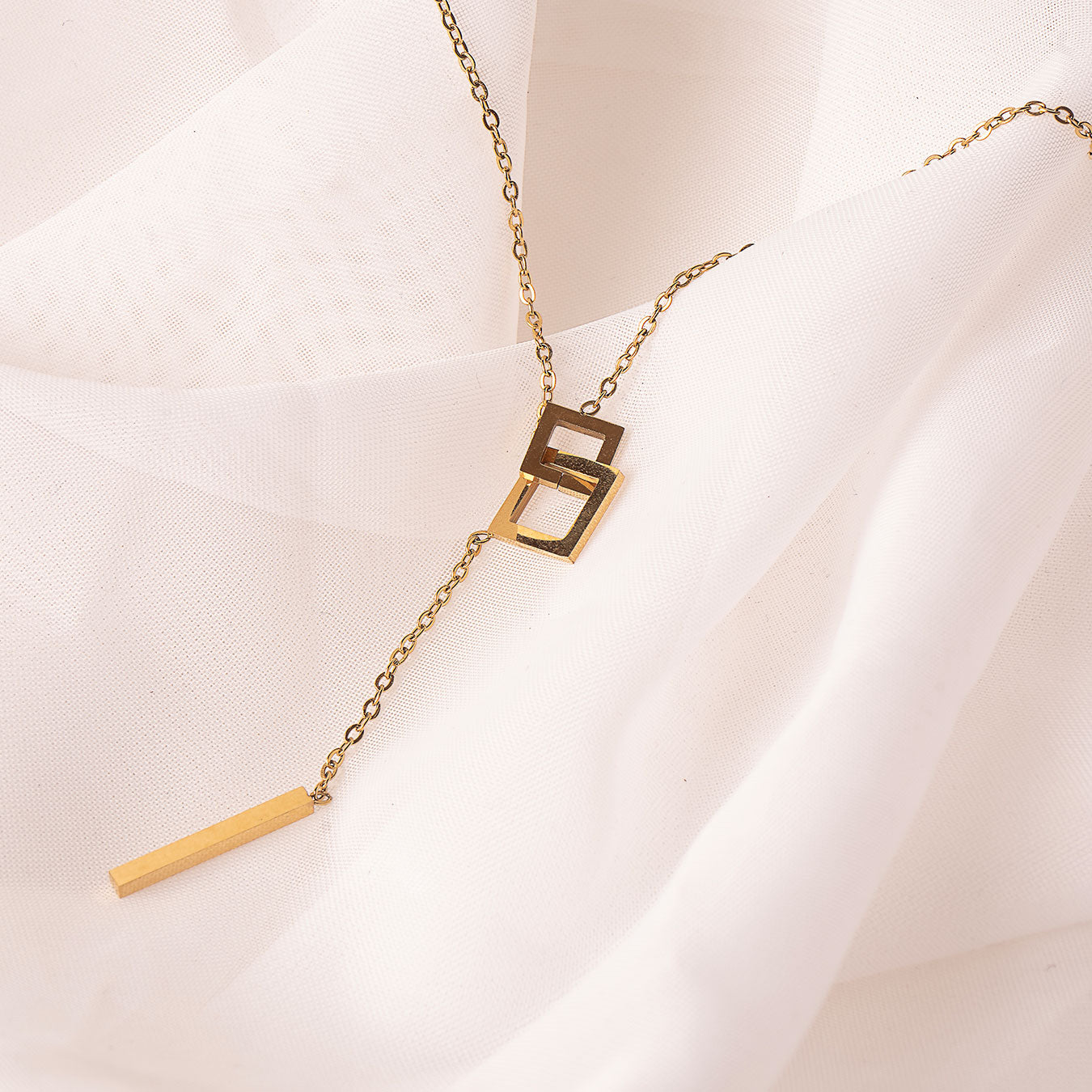 A striking 18k gold plated Jasmine Grace Necklace, featuring a modern lariat style with an interlocking square detail and a sleek rectangular bar pendant, elegantly displayed on a white textured fabric.