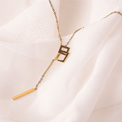 A striking 18k gold plated Jasmine Grace Necklace, featuring a modern lariat style with an interlocking square detail and a sleek rectangular bar pendant, elegantly displayed on a white textured fabric.