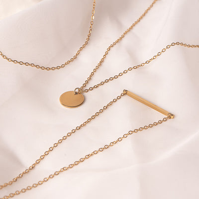 Three elegant 18k gold plated Camellia Classic Chain necklaces are tastefully arranged on a soft, draped white fabric. One features a sleek horizontal bar pendant, another a simple circular disc pendant, and the third is a delicate plain chain, showcasing minimalist style.