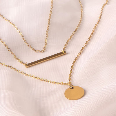 Three elegant 18k gold plated Camellia Classic Chain necklaces are tastefully layered on a soft, draped white fabric. One features a sleek horizontal bar pendant, another a simple circular disc pendant, and the top chain is a delicate plain link, showcasing minimalist style.