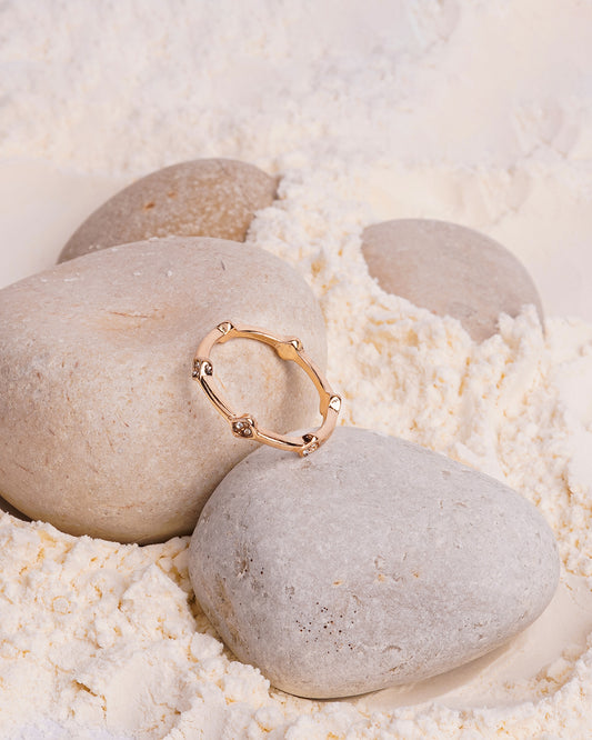 The Amara Bamboo Ring is showcased resting on a smooth white stone, surrounded by other stones and a bed of white powder. The gold-toned ring has a segmented, bamboo-like design, with a few of the segments accented by small, clear gemstones.