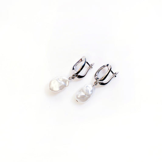 A top-down studio shot against a white background shows a pair of Alba Teardrop Pearl Drops earrings. Each silver-toned earring features a polished, tear-drop shaped loop from which a single, lustrous, irregularly shaped white baroque pearl hangs.