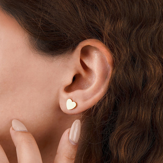 A close-up side profile of a person's ear, wearing a single Amara Heart Stud. The small, polished gold heart earring rests elegantly on the earlobe, complementing the person's clean nail polish and soft hairstyle.
