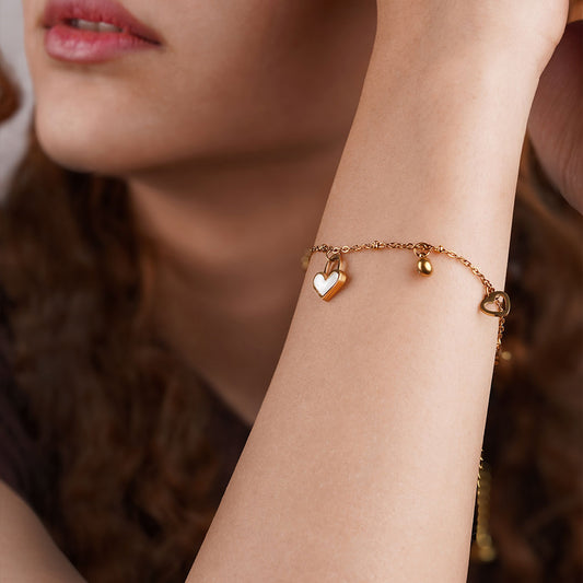 A close-up shot of a person's wrist wearing the Amara Love Charms bracelet. This delicate gold-toned chain bracelet is adorned with several charms, including a small, white heart charm and a minimalist open heart charm, resting on the top of the wrist.