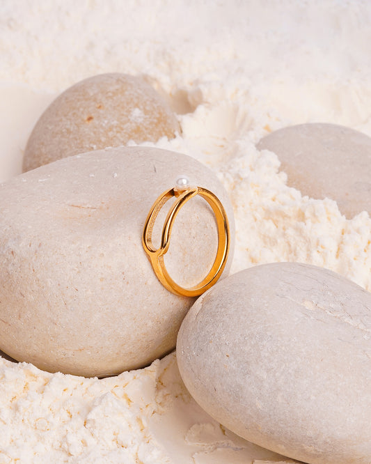 The Anna Pearl Duo Ring is showcased resting on a smooth white stone, surrounded by other stones and a bed of fine white powder. The gold-toned open ring's double bands elegantly frame a single, polished white pearl.