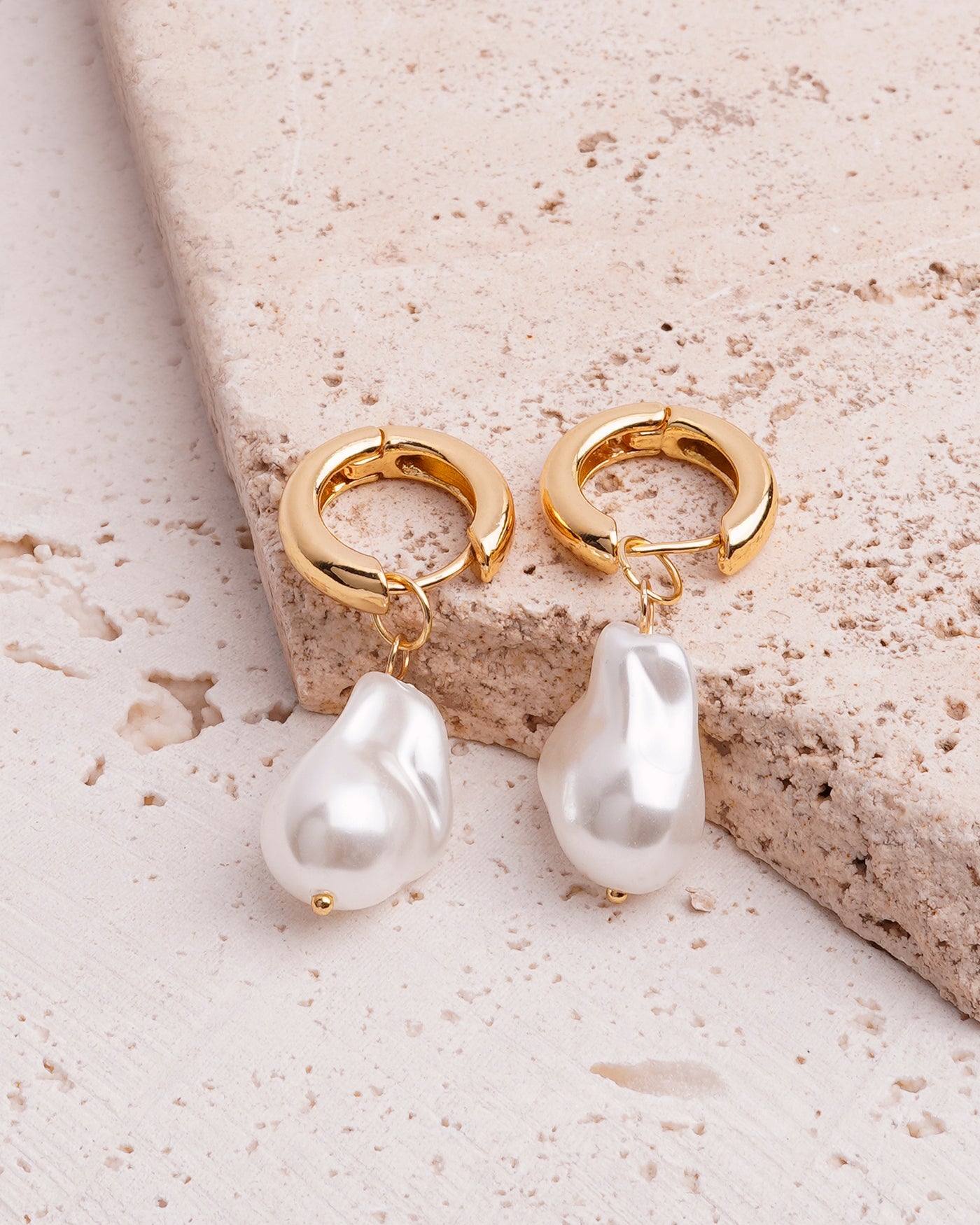 The Aurelia Pearl Drop Hoops are showcased on a beige, textured stone surface. The gold-toned hoops are connected to large, lustrous white baroque pearls, with one earring propped up to highlight its unique shape.