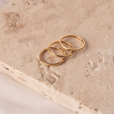 Aurora Shimmer Ring by Delta Jewel, a minimalist gold-plated band with a subtle sparkle finish, shown stacked with two plain bands on a textured stone surface.