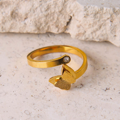 The Aurora Butterfly Ring is displayed on a textured stone surface. This gold-toned open ring has a small, brilliant gemstone on one end and a beautifully detailed butterfly with a textured wing on the other, creating an elegant and artistic design.