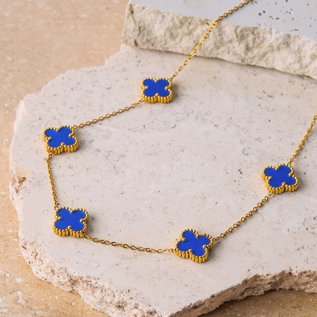  The Aurora Clover Pendant is elegantly draped over a textured, light-colored stone surface. This gold necklace showcases five vibrant blue clover-shaped pendants evenly spaced along its delicate chain.