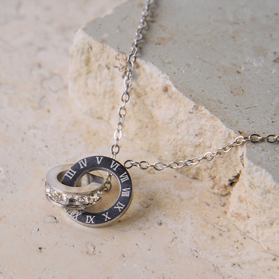 The Aurora Trend Necklace is displayed on a beige, textured stone surface. This silver-toned necklace features a delicate chain connected to a centerpiece of two interlocking rings: one with Roman numerals and the other with a band of sparkling cubic zirconia.