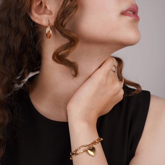 A close-up side profile of a person's head and arm, showcasing multiple gold-toned jewelry pieces. A thick, textured hoop earring dangles from the ear, and a delicate chain bracelet with a single heart charm is on the wrist. A simple, thin open ring is also visible on the person's finger.
